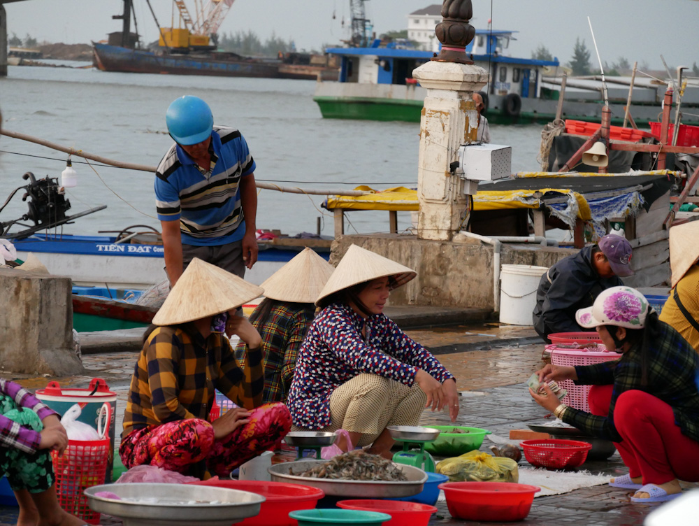 Vendors at Ha Tien's morning fish market, with fishing trawlers in the background as tehy travel up the Dong Ho estuary. 